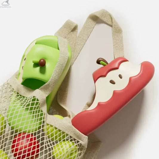 Red and green Apple Cloud Slides tucked into a mesh market bag next to fresh apples, featuring cute cloudsharks cushion comfort design.