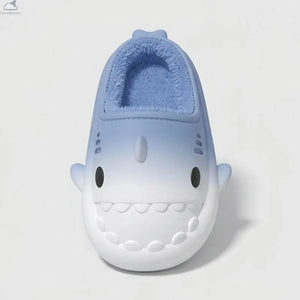 A single children's slip-on shoe shaped like a smiling shark, shown from the front on a clean light gray studio background. The upper is light blue with a white lower section, black eye details, and a fuzzy blue lining visible at the opening.